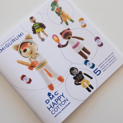 Livro capa branca com bonecos de crochê coloridos e textos AMIGURUMI, DMC HAPPY COTTON e número 5