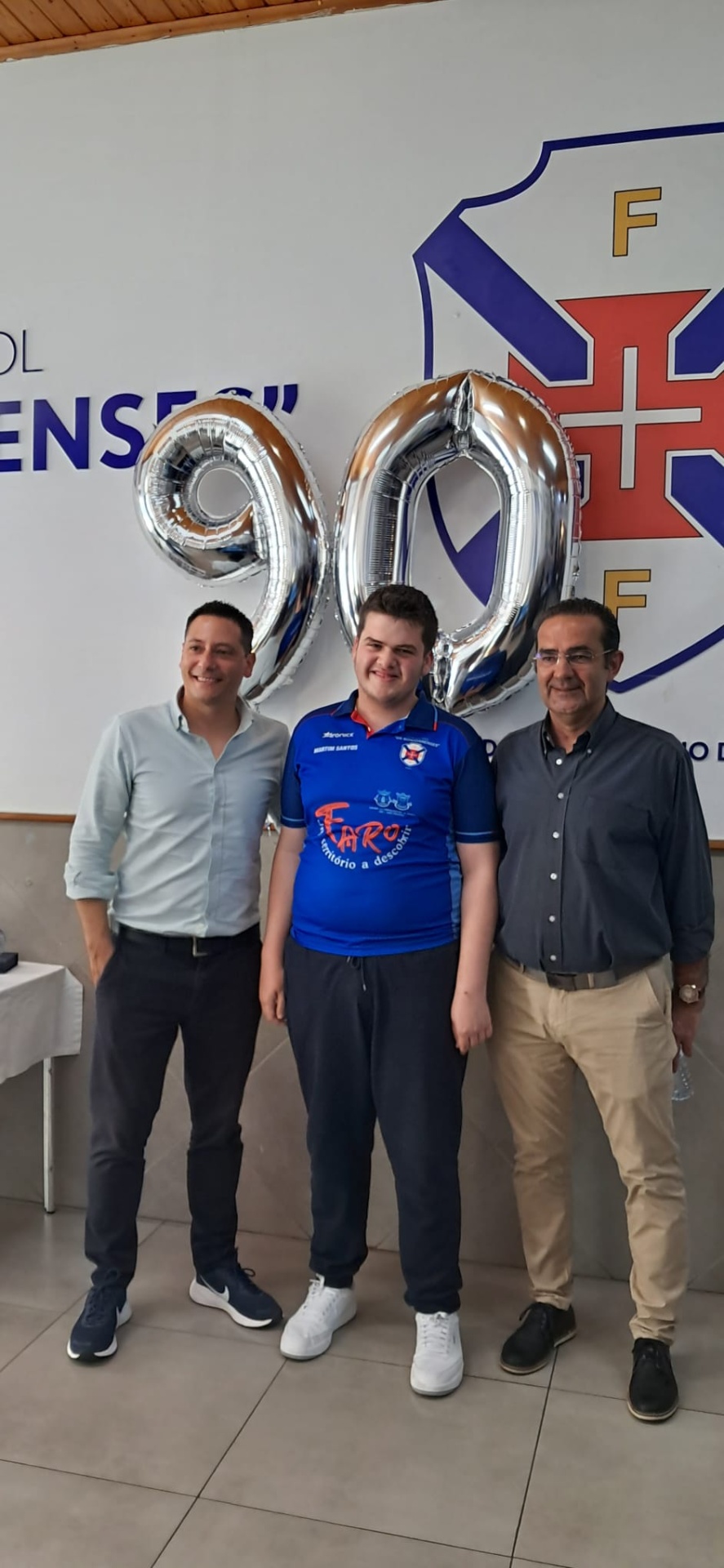 Três homens em pé com balões prateados em forma de números 9 e 0 e um emblema colorido no fundo