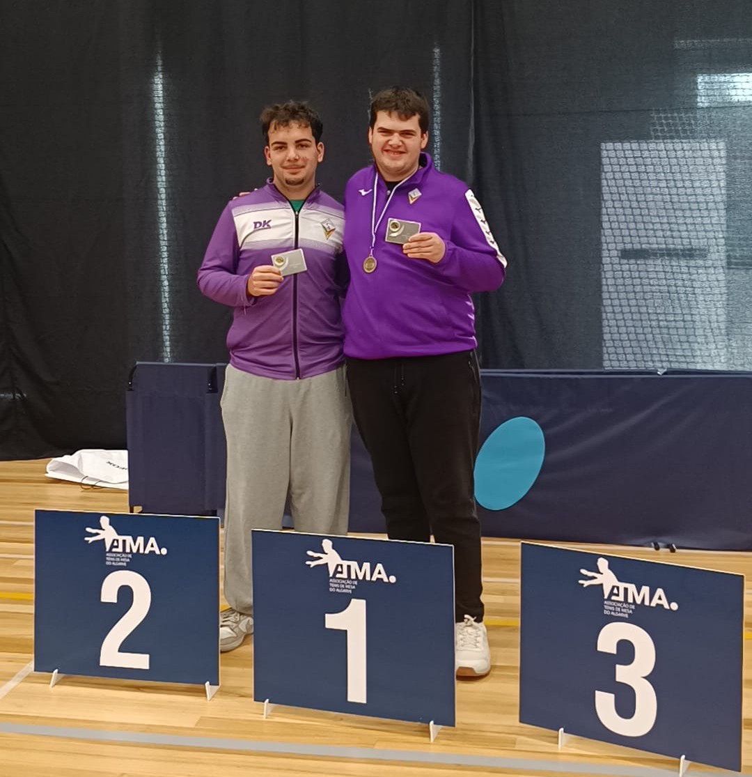 Dois homens com casacos desportivos roxos seguram prémios em pista com sinais de podium 1, 2, 3.