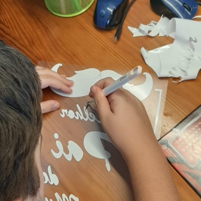 Pessoa a aplicar decalque branco com texto em suporte transparente numa mesa de madeira com materiais de escrita e rato por perto