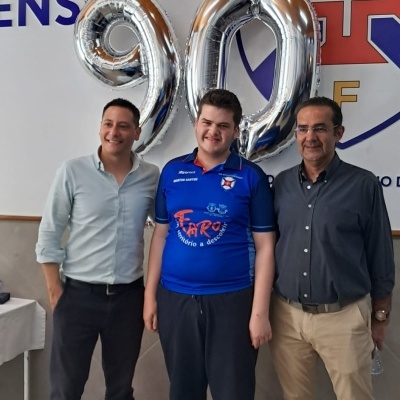 Três homens em pé com balões prateados em forma de números 9 e 0 e um emblema colorido no fundo