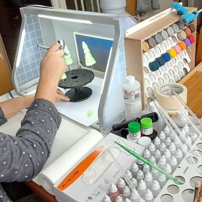 Pessoa a usar aerógrafo para pintar miniaturas verdes numa cabina de pintura com tintas e acessórios para modelismo