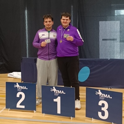 Dois homens com casacos desportivos roxos seguram prémios em pista com sinais de podium 1, 2, 3.