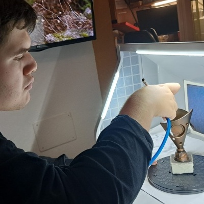 Pessoa detalhando objeto metálico triangular dentro de caixa branca iluminada.