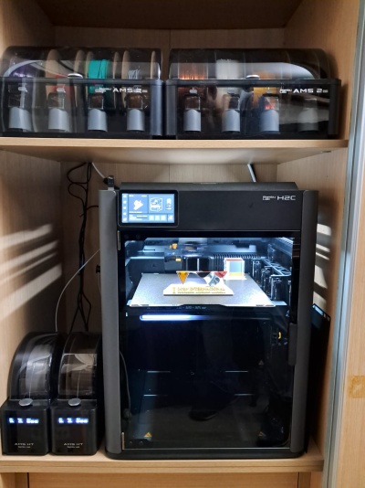 Impressora 3D preta Bambu Lab H2C em estante de madeira com dispensadores de filamento.