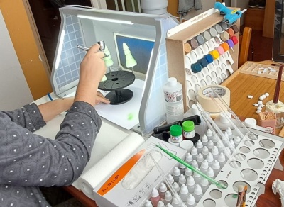 Pessoa a usar aerógrafo para pintar miniaturas verdes numa cabina de pintura com tintas e acessórios para modelismo