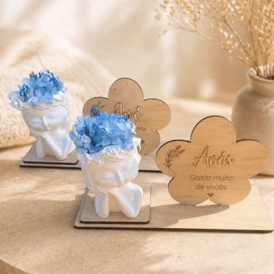 Esculturas brancas de rostos sorridentes com flores azuis e placas de madeira com texto em português sobre uma superfície clara.
