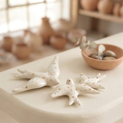 Pássaros decorativos em cerâmica branca com manchas douradas sobre mesa clara, ao lado de tigela de barro com pedras e folhas secas.