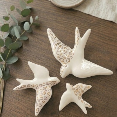 Três pássaros decorativos em cerâmica branca com detalhes dourados sobre mesa de madeira.