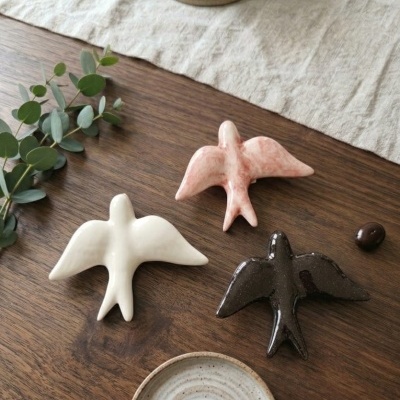 Três esculturas decorativas de pássaros em cerâmica nas cores branco, rosa e preto sobre mesa de madeira