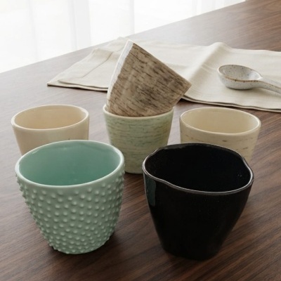 Conjunto de copos de cerâmica em diferentes cores e texturas sobre mesa de madeira com pano de mesa e colher.