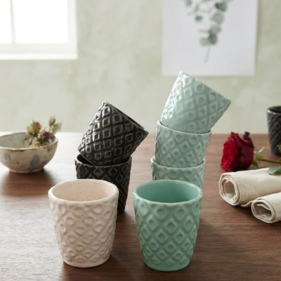 Copos de cerâmica texturizados em creme, azul claro e preto sobre mesa de madeira com guardanapos e rosa vermelha.