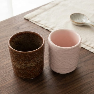 Dois copos cerâmicos, castanho e rosa, sobre mesa de madeira com toalha creme e colher cerâmica