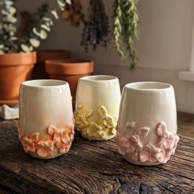 Três vasos de cerâmica creme com flores em relevo coloridas sobre mesa de madeira rústica