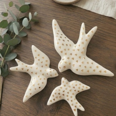 Três pássaros decorativos brancos com corações dourados sobre mesa de madeira com folhas verdes e peça cerâmica.