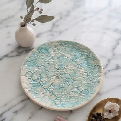Prato decorativo azul e branco em cerâmica com padrão texturizado, vaso branco com folhas secas, pires dourado com pedras e grãos de café, sobre superfície marmoreada.