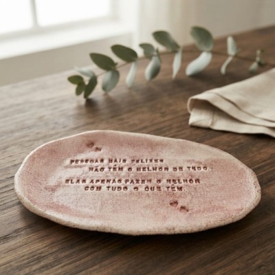 Prato cerâmico rosa com texto gravado em relevo, colocado numa mesa de madeira com planta e pano ao lado