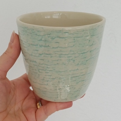 Caneca de cerâmica creme com texturas azuis clara, segurada por mão em fundo branco