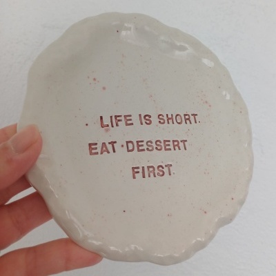 Prato de cerâmica bege com texto em vermelho e borda irregular, segurado à mão