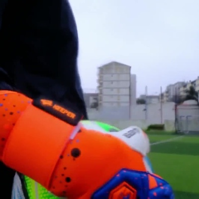 Luvas de guarda-redes laranja e azul usadas por jogador num campo de futebol