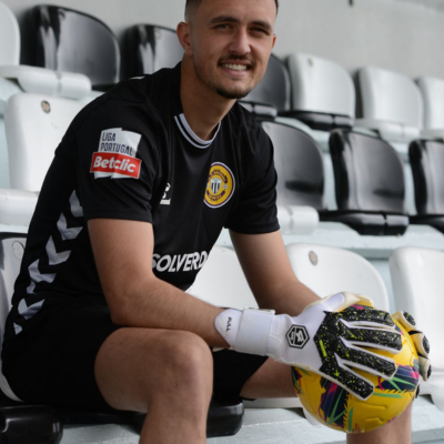 Jogador de futebol sentado com camisola preta e luvas, segurando bola amarela colorida em bancadas do estádio