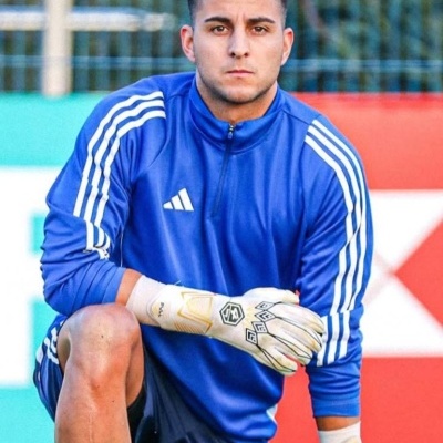 Homem com camisola azul Adidas e luvas de guarda-redes a ajoelhar no campo