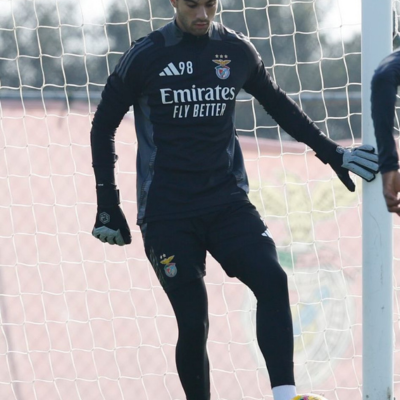 Jogador de futebol com roupa preta de treino, luvas e bola amarela perto da baliza