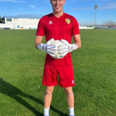 Jogador de futebol com uniforme vermelho e luvas brancas segurando bola no campo.