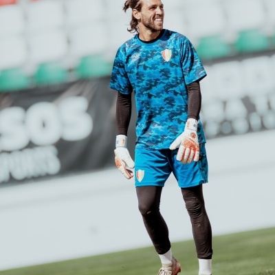 Jogador de futebol com equipamento azul camuflado, luvas brancas e calções azuis no campo