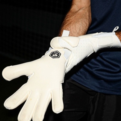 Luvas de guarda-redes brancas com logótipo preto e branco e detalhes em velcro, usadas por pessoa com roupa azul e preta.