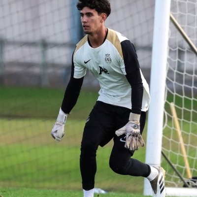 Guarda-redes com equipamento branco, preto e dourado a defender baliza no campo de futebol
