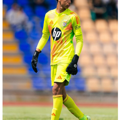 Guarda-redes de futebol com uniforme amarelo fluorescente, logos HP e Adidas, luvas pretas e chuteiras cinza e verde em campo de relvado.