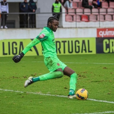 Jogador de futebol em campo vestindo kit completo verde claro a pontapear bola amarela