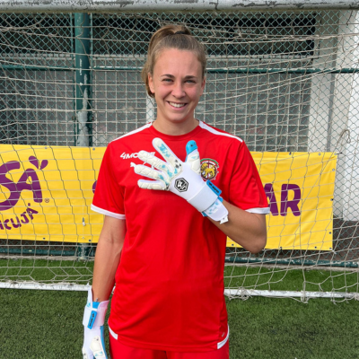 Mulher com equipamento vermelho e luvas de guarda-redes em campo de futebol diante da baliza