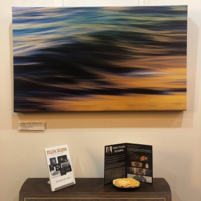 Pintura abstrata colorida pendurada numa parede branca sobre uma mesa com livros e um prato.