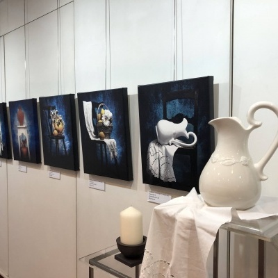 Seis pinturas de naturezas-mortas penduradas numa parede branca e jarro branco de cerâmica sobre mesa com pano branco