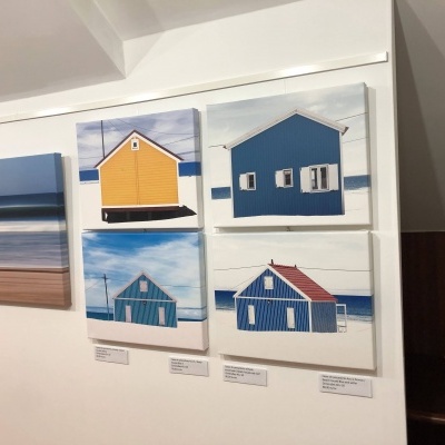 Quadros com imagens de casas de praia em madeira coloridas expostos numa galeria