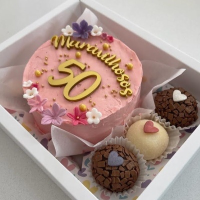 Bolo rosa com decoração floral e brigadeiros numa caixa branca.