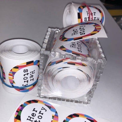 Rolos de autocolantes com 'Her story' em branco e padrão colorido numa caixa transparente
