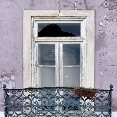 Janela antiga com vidro partido e varanda de ferro forjado preto numa parede lilás gasta