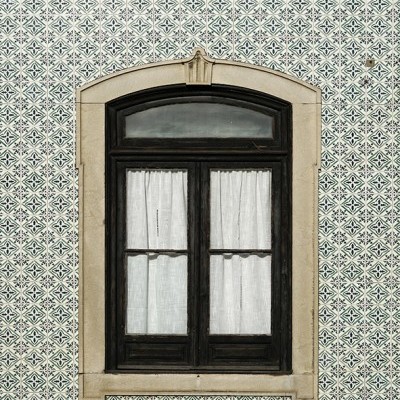 Janela de madeira preta com cortinas brancas em parede de azulejos azuis e brancos