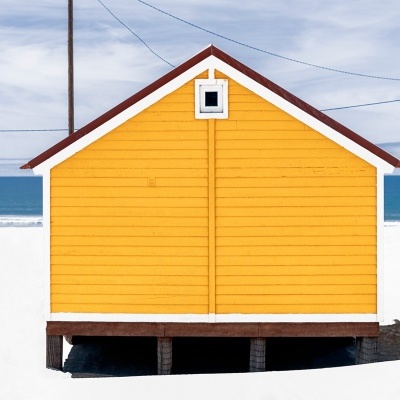Casa pequena de praia amarela sobre pilares na areia com mar ao fundo
