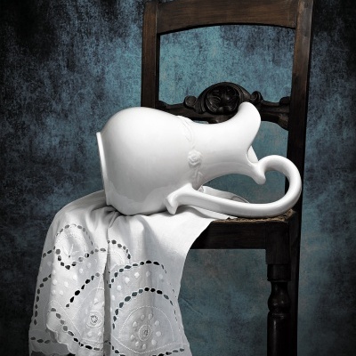 Jarra branca de cerâmica sobre toalha branca bordada em cadeira de madeira escura.