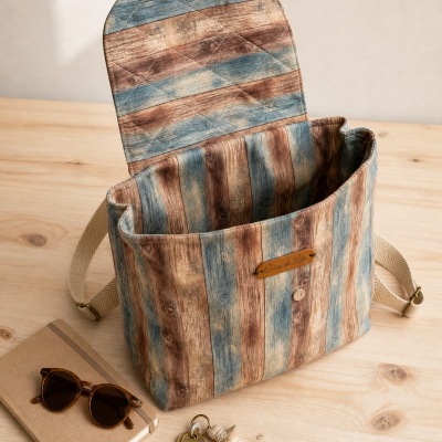 Mochila com padrão de madeira envelhecida em tons azuis, castanho e bege, com etiqueta de couro Coisas da Lika