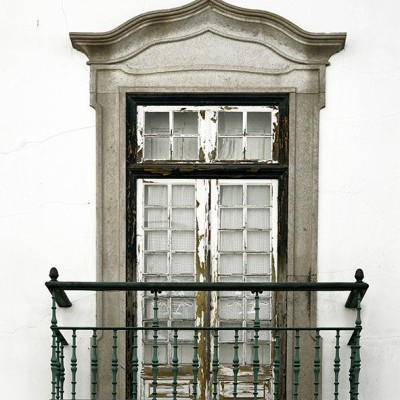 Janela de madeira preta desgastada com varanda de ferro verde numa parede branca