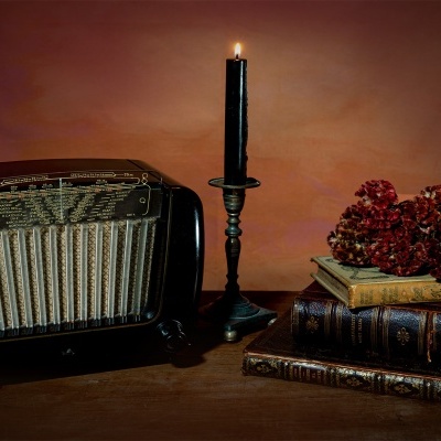 Rádio antigo, vela preta acesa num castiçal e livros antigos com flores secas numa mesa de madeira