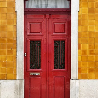 Porta dupla vermelha em madeira com vidros e moldura branca numa parede com azulejos amarelos e número 196