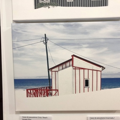 Tela com imagem de casa de pescadores branca e vermelha junto ao mar sob céu nublado.