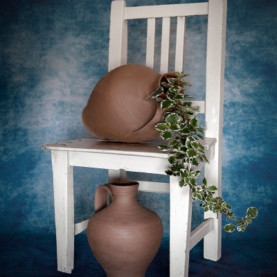 Cadeira branca envelhecida com vaso de barro e planta, jarra de barro no chão, fundo azul.