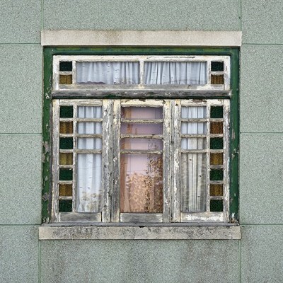 Janela antiga de madeira com vidro colorido e cortinas brancas numa parede verde com base cinzenta